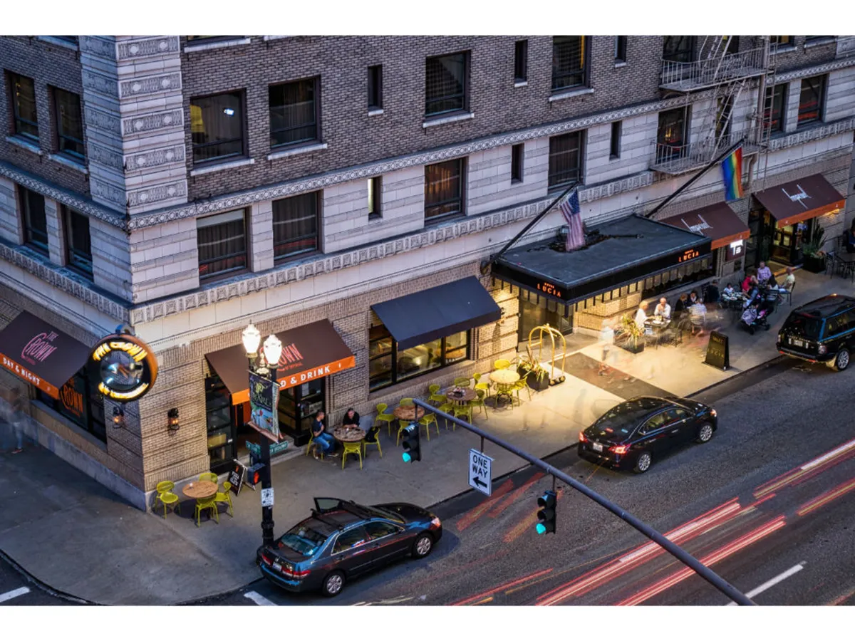 A colorful restaurant patio scene with bright green chairs, an illuminated sign, and people dining outdoors. The patio is alive with conversation and energy, set against the backdrop of a busy city street signaling the social and cultural pulse of the neighborhood.