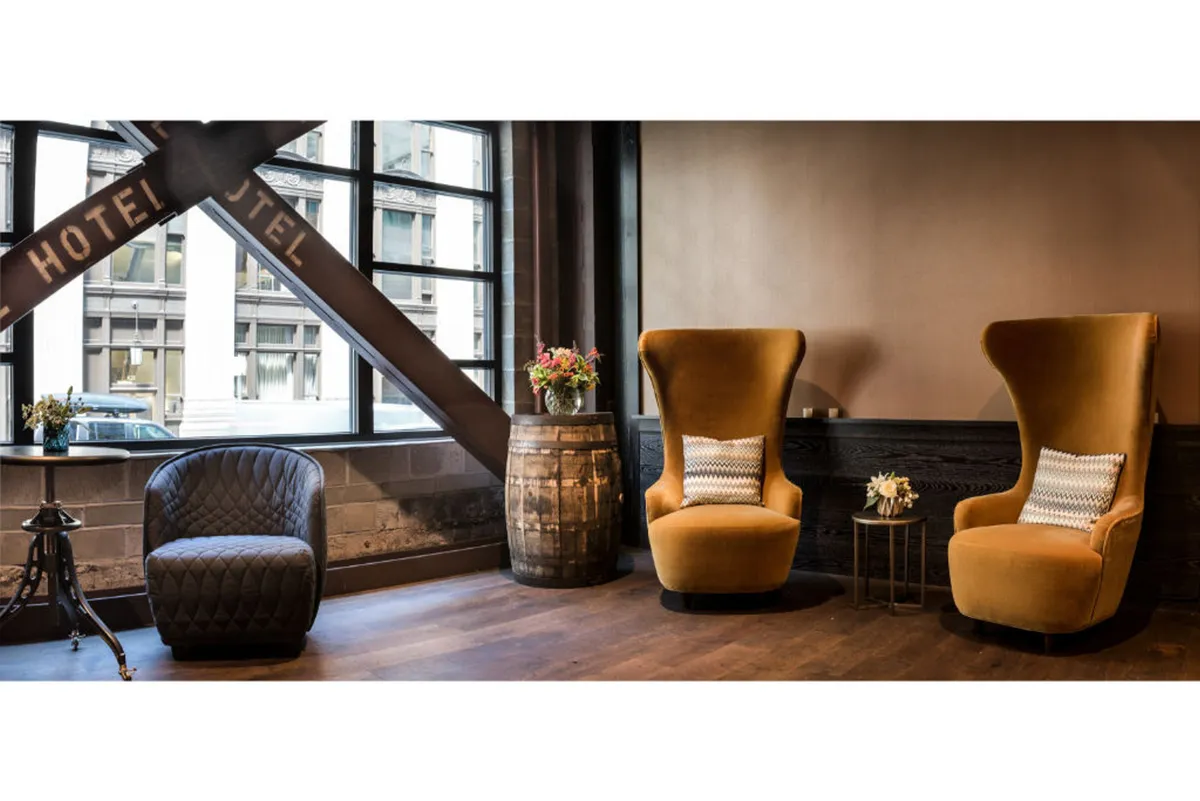 A cozy nook with two large yellow chairs, a black chair, a wooden barrel table, and a fresh floral arrangement near bright windows, inviting guests to unwind and people-watch.