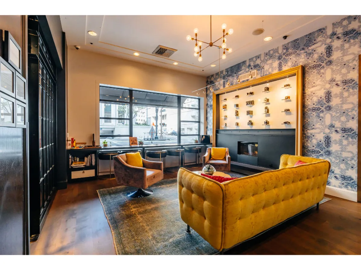 A stylish lounge area featuring a yellow tufted sofa, chic armchairs, a display shelf with local decor, and expansive windows creating a bright, welcoming atmosphere.