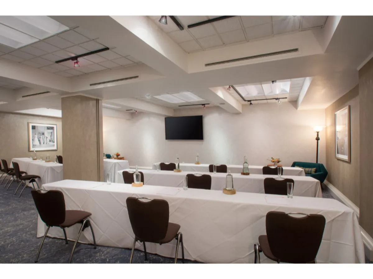 A long table dressed in crisp white tablecloths surrounded by comfortable brown chairs, with a television screen mounted on the wall and a decorative lamp illuminating the corner. The setting is ideal for focused meetings, collaborative sessions, or creative business presentations.