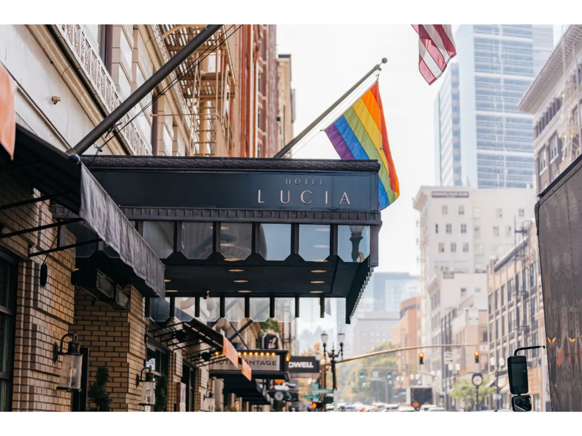A vibrant exterior shot of the hotel with the prominent 'Hotel Lucia' signage, rainbow and American flags, and bustling street activity in the foreground. The scene captures a welcoming atmosphere at the threshold between hotel and city.