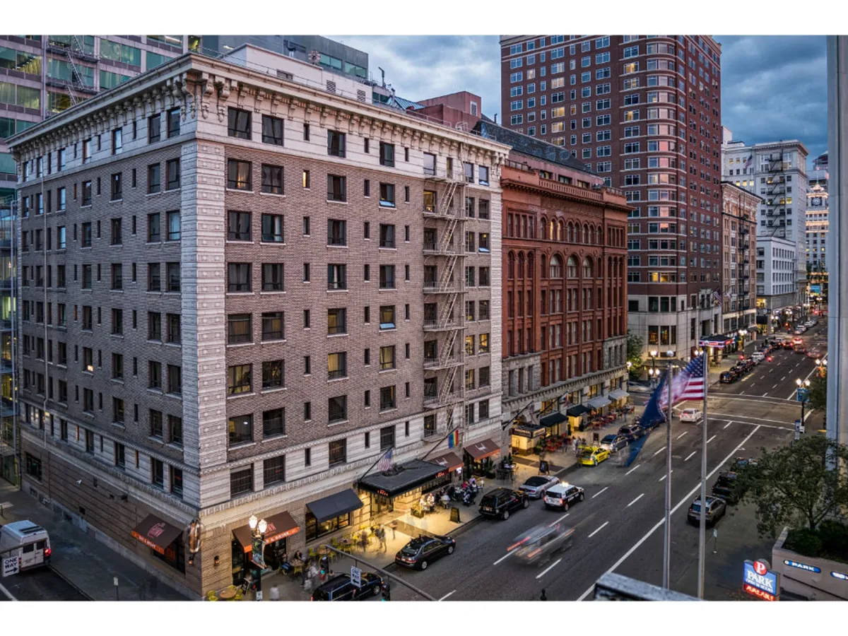A lively city street scene featuring historic Portland buildings, bustling sidewalk activity, and flags waving alongside passing vehicles. The architecture reflects the heritage of downtown Portland, with people engaged in shopping, dining, or heading to cultural events in the area.