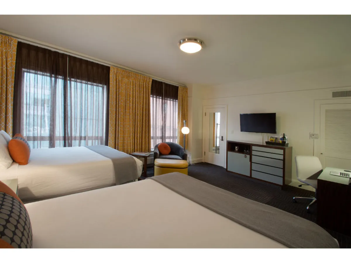 The image showcases a stylish hotel room with two inviting beds, a cozy seating area featuring an armchair, a sleek TV unit, and expansive windows draped in soft curtains that let in abundant natural light. The space feels warm, comfortable, and thoughtfully designed for both relaxation and productivity, with a desk positioned to enjoy views of downtown Portland.