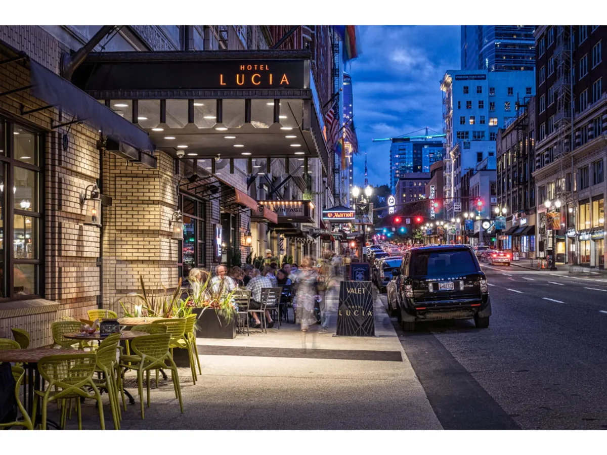 The illuminated hotel entrance with outdoor seating, people enjoying al fresco dining, and lively street activity at dusk. The scene blends the comfort of the hotel with the energy of Portland’s urban lifestyle.
