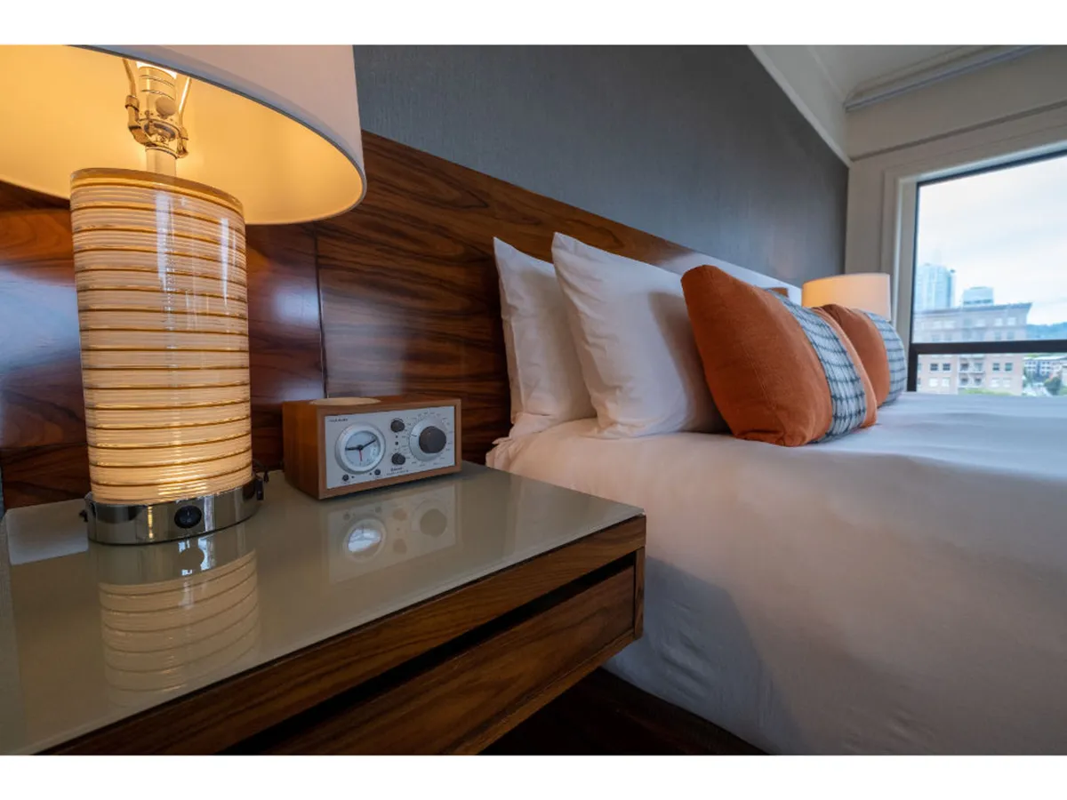 A plush bed with accent pillows is flanked by a nightstand lamp and a retro-style clock radio. The space feels cozy and serene, illuminated softly by natural light. The details create an atmosphere of restful retreat, ideal for relaxation or quietly reading before bed.