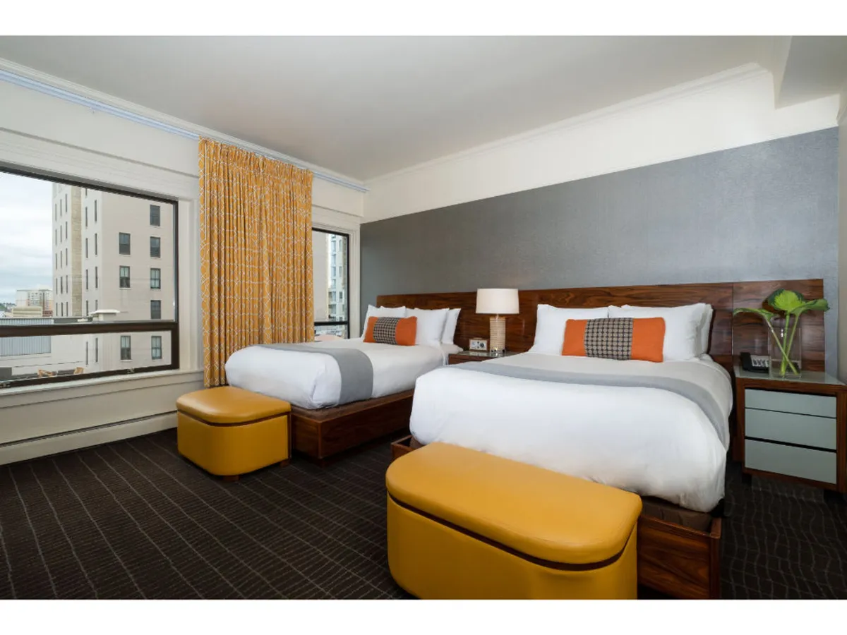 This image features two beds with crisp white linens, decorative pillows, and a yellow ottoman at the foot. Large windows let in abundant natural light, complemented by bold orange curtains that add a modern pop of color. The overall mood is contemporary yet comfortable, with an inviting layout that encourages both relaxation and socializing.