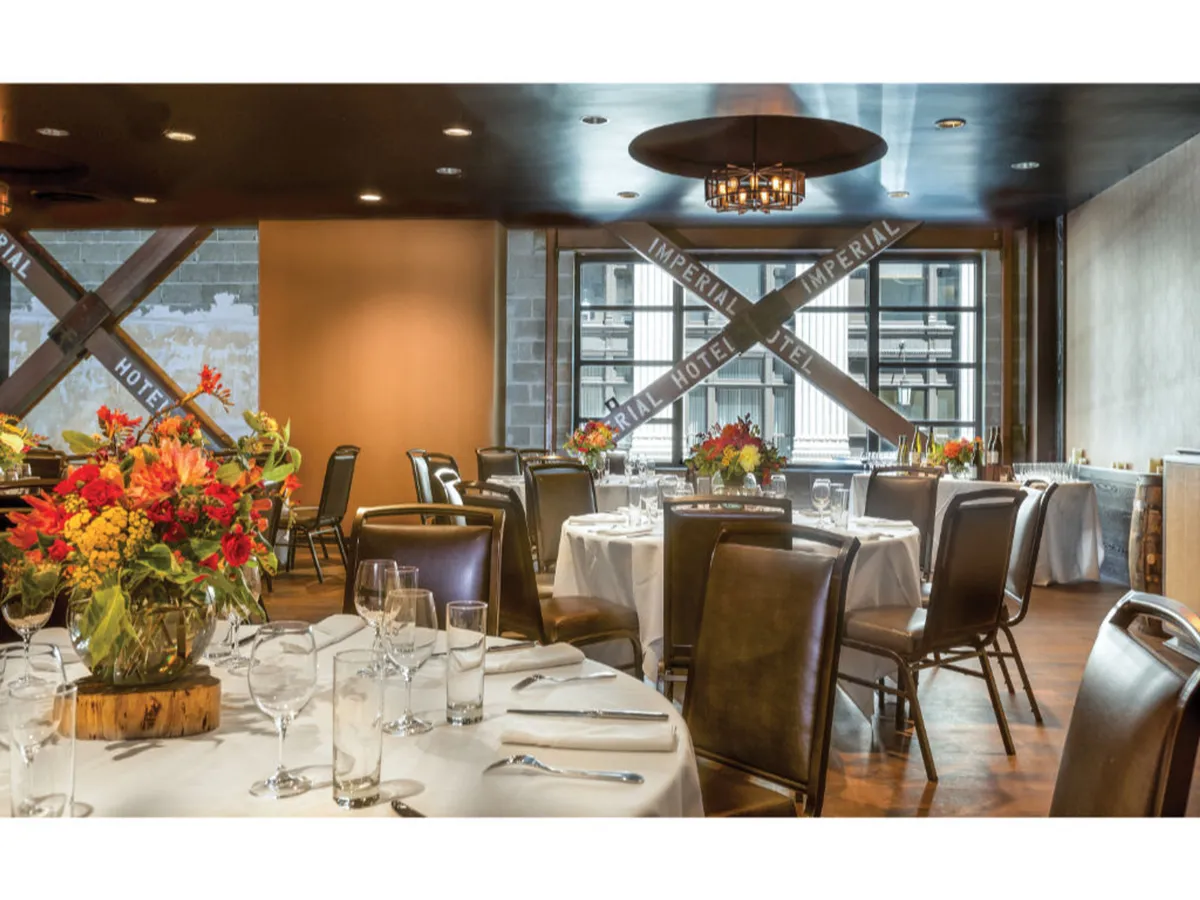 This image presents an elegant dining room with large windows offering sweeping city views, set for a sophisticated meal with decorative floral arrangements and polished glassware.